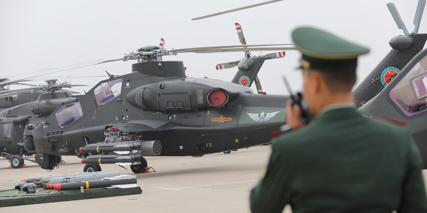 Soldat spricht in Funkgerät, während im Hintergrund Kampfhubschrauber stehen.