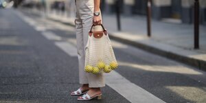 Eine Frau steht auf der Straße, in ihrer Hand eine Fischnetztasche, durch die man gelbe Äpfel sehen kann.