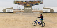 Ein kleiner Junge sitzt auf seinem Fahrrad, im Hintergrund ragt wuchtig das Gründungsdenkmal der Weltraumstadt Baikonur empor.
