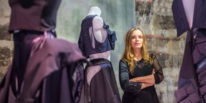 Modedesignerin Nina Holleins steht im Ausstellungsraum neben ihrer Kollektion von violetten Kleidern vor einer großem Leinwand