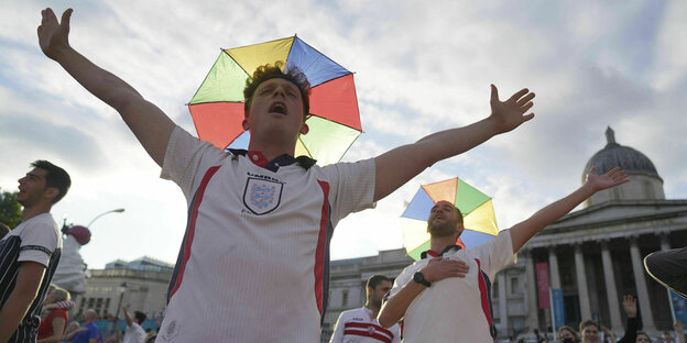Ein Fussballfan mit ausgebreiteten Armen und Regenbogen-Schirm als Kopfbedeckung