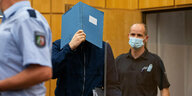 Der Hauptangeklagte im Fall Münster wird in den Gerichtsaal geführt und hält sich einen blauen Ordner vors Gesicht