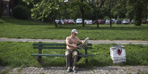 Ein älterer Mann mit nacktem Oberkörper sitzt auf einer Parkbank und liest eine Zeitung