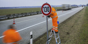 Ein Mann mit oranger Arbeitskleidung steht auf einer Leiter und montiert ein Tempo-130-Schild