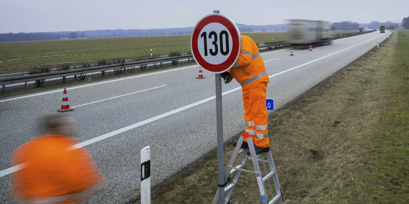 Ein Mann mit oranger Arbeitskleidung steht auf einer Leiter und montiert ein Tempo-130-Schild