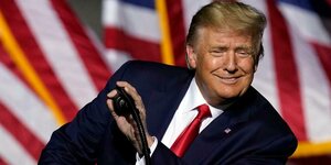 Donald Trump steht vor einem Rednerpult und hält seine Hand über das Mikrophon