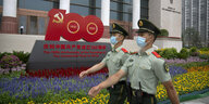 Cinesische Polizisten in Uniform mit Mundschutz laufen vor einem Gebäude uned einem Blumenbeet mit Logo 100 Jahre kommunistische Partei vorbei