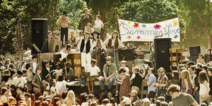 Eine Hippie-Band spielt auf einem Lastwagen, umringt von vielen Menschen. Im Hintergrund ist ein Transparent zu sehen, auf dem steht: "Summer of Love"