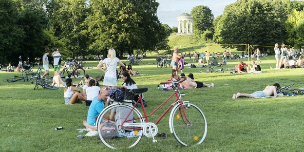 Eine Liegewiese des Englischen Gartens in München. Im Vordergrund ein Fahrrad, im Hintergrund Menschengruppen die am Boden sitzen oder liegen.