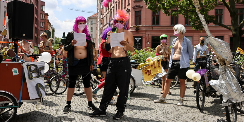 Eine Gruppe von Demonstrierenden steht in der Sonne auf einem Platz. Manche von ihnen haben Mikros in der Hand und halten eine Rede während der Kundgebung. Sie sind alle oben ohne, tragen dafür aber Perücken und Masken.