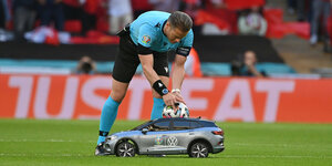Ein Fußballschiedsrichter beugt sich über ein kleines Elektroauto, um den Ball zu greifen, der auf dessen Dach liegt