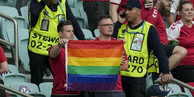 Zwei Fans stehen im Stadion und halten eine Regenbogenflagge, flankiert von Ordnern