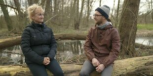 Rechts sitzt Richard Zschech, links von ihm seine Mutter. Sie sitzen auf einem Baumstamm und tragen Herbstjacken. Hinter ihnen ist ein Teich und Bäume ohne Blätter. Zschech lächelt, seine Mutter runzelt die Stirn.