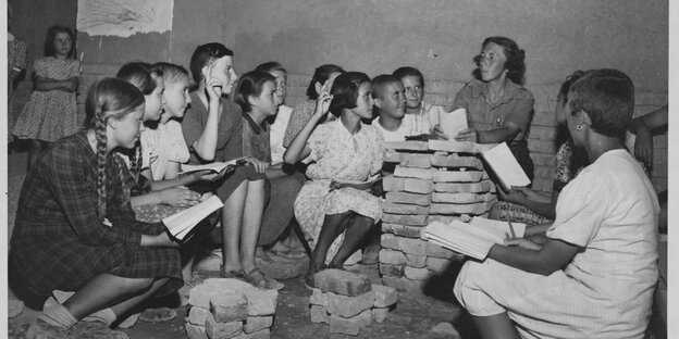 Eine schwarzweiße Fotografie aus den 1940er Jahren, Kinder sitzen auf gestapelten Ziegeln, Bücher auf den Knien.