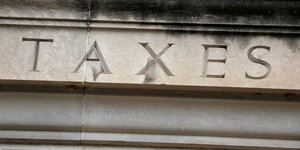 Auf einer massiven Steinwand ist in einem Bildausschnitt die eingravierte Aufschrift "Taxes" zu lesen
