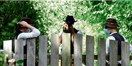 Drei Männer mit altertümlichen Hüten und Westen stehen hinter einem Zaun. Einer fotografiert nach draußen.