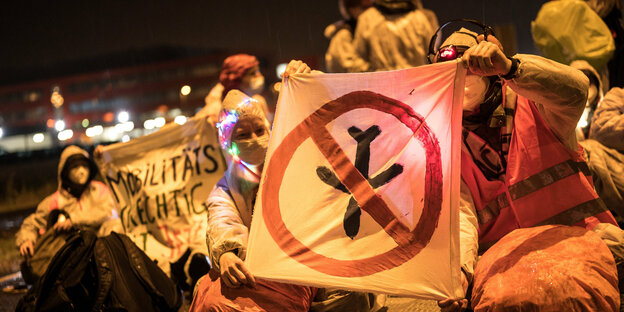 Demonstrierende sitzen auf einer Straße und halten ein Banner mit durchgestrichenem Flugzeug