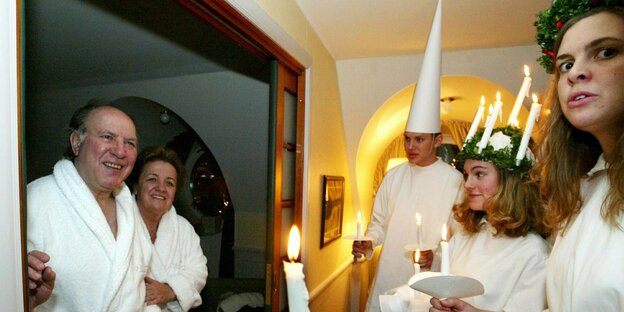 Zwei Menschen in Bademänteln werden von weißgekleideten Menschen mit brennenden Kerzen am schwedischen Feiertag Lucia-Tag begrüßt