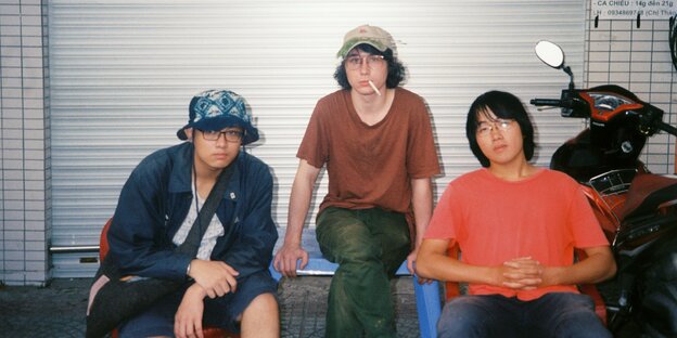 Die drei Musiker sitzen vor einer Garage, daneben ein Motorroller