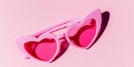 Auf einem pinken Untergrund liegt eine pinke Brille, deren Gläser herzförmig und rot gefärbt sind