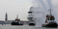 Kreuzfahrtschiff mit 3 Lotsenbooten, die es mit Wasserfontänen begrüßen, im Hintergrund Venedig