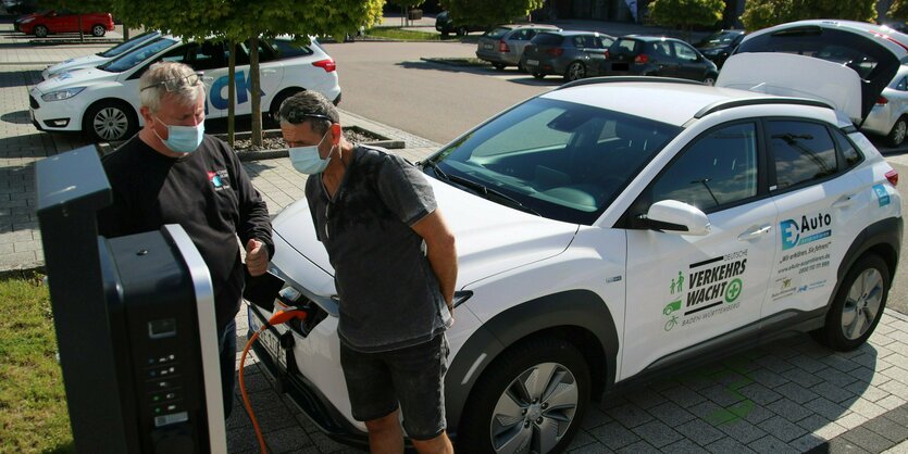 Zwei Männer mit Mundschutz stehen vor einer Ladesäule - hinter ihnen ein Elektroauto