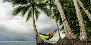 Eine Person liegt in einer Hängematte, die an Palmen mit Blick auf das Meer festgezurrt ist