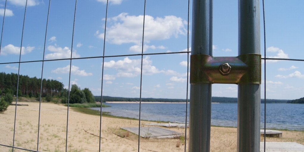 Ein Bauzaun versperrt den Weg zum Sandstrand am Helenesee bei Frankfurt (Oder). Der Himmel ist blau.