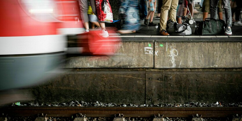 Ein Zug fährt in den Bahnhof ein, Menschen stehen am Bahnsteig