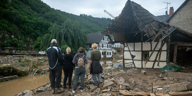 4 Menschen stehen mitd em Rücken zur Kamera in einem vom Hochwasser zerstörten Dorf, ein Fachwerkhaus und gelber Schlamm umgibt sie