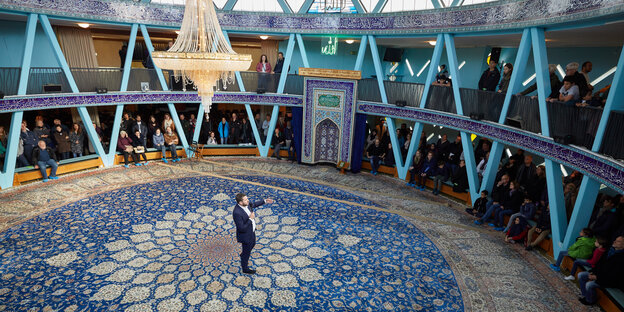 Ein Mann steht in der Mitte eines Raumes mit blauen Ornamenten auf dem Boden und einem großen Kronenleuchter an der Decke. Ringsum sitzen Menschen am Rand des runden Raumes