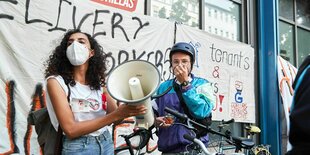 Eine Frau mit Megafon und ein Mann mit Fahrradhelm und Türkiser Jacke stehen vor einer Häuserfassade, an der ein Transparent festgemacht ist