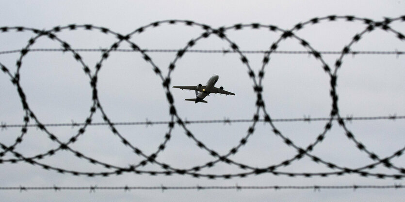 Blick durch Stacheldraht auf ein Flugzeug am Himmel