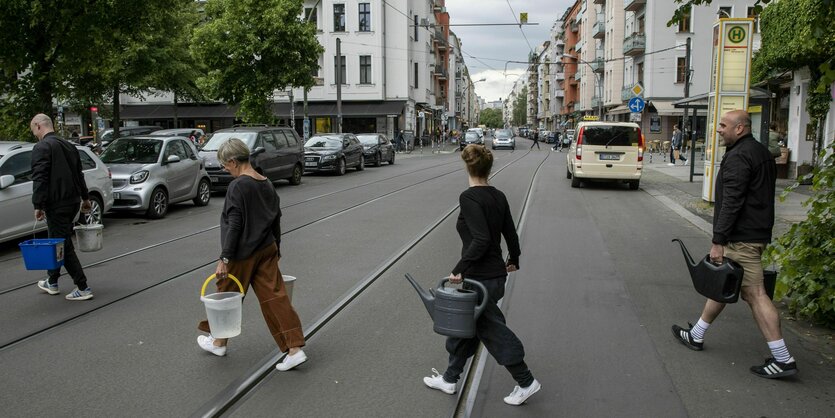 Vier Menschen gehen über eine Straße und tragen Gießkannen und Eimer voll Wasser.