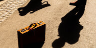 Ein altmodischer Koffer, ohne Rollen und Schischi, steht auf einem Weg. lange Schatten von zwei Personen umgeben den Koffer.