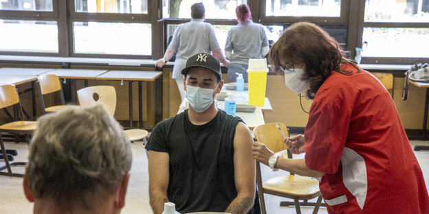 In einem Klassenraum sitzt ein Schüler mit medizinischer Maske der von einer Frau geimpft wird.
