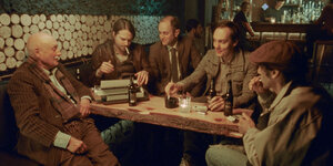 In einer Kneipe sitzen fünf mittelalte Männer um einen Tisch herum