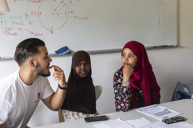 Ein Mann spricht einen Buchstaben laut aus und zeigt dabei auf die Spitze seiner Zunge, zwei Mädchen schauen ihm dabei genau zu: Sprachunterricht mitten in den Sommerferien im Nachbarschaftshaus in Berlin-Neukölln