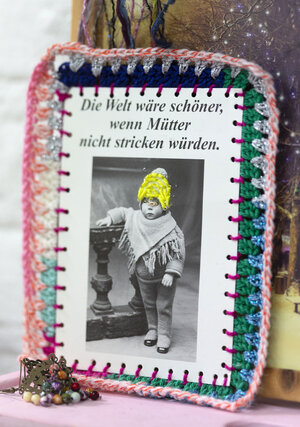Eine mit Garn verzierte Postkarte, auf der steht: Die Welt wäre besser, wenn Mütter nicht stricken müssten Eine mit Garn verzierte Postkarte, auf der steht: Die Welt wäre besser, wenn Mütter nicht stricken müssten