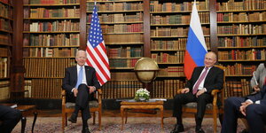 Zwei Männer sitzen vor einer Wand mit alten Büchern sowie einer amerikanischen und russischen Flagge nebeneinander, zwischen ihnen ein Globus