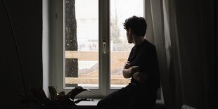 Eine Person schaut aus dem Fenster. Man sieht nur Schattierungen und dass die Person die Arme verschränkt hält