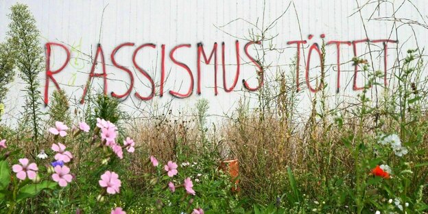 An einer Wand ist der rote Schriftzug "Rassimus tötet" gesprayt, davor wachsen Blumen auf einer Wiese