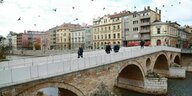 Menschen gehen über eine Brücke mit einer Altstadt im Hintergrund und umher fliegenden Vögeln