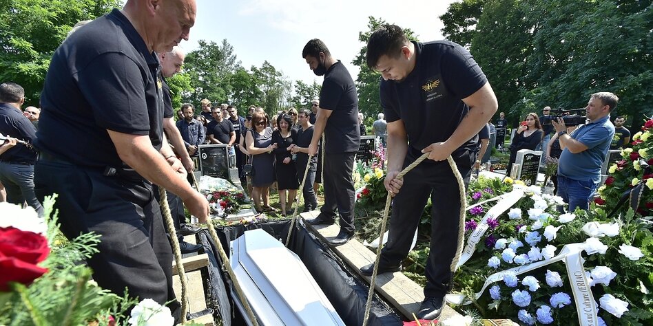 Vier Männer lassen einen Sarg in ein Grab hinab. Um sie herum stehen viele Trauergäste