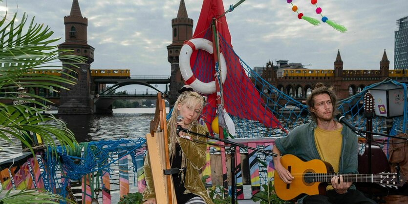 Zwei Musiker:innen spielen auf einem Boot. Im Hintergrund ist die Oberbaumbrücke