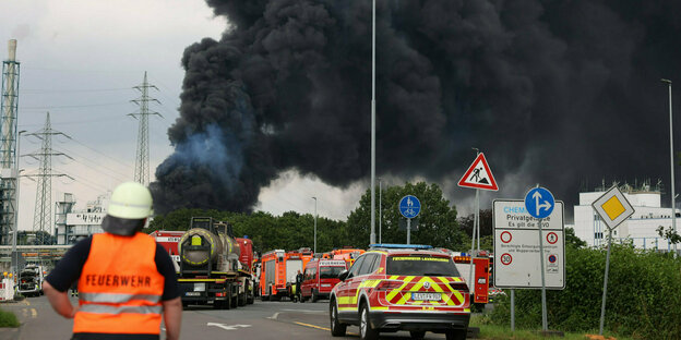 Leverkusen: Einsatzfahrzeuge der Feuerwehr stehen unweit einer Zufahrt zum Chempark über dem eine dunkle Rauchwolke aufsteigt.