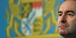 Gesicht von Hubert Aiwanger rechts im Bild, dahinter unscharf das Wappen des Freistaats Bayern