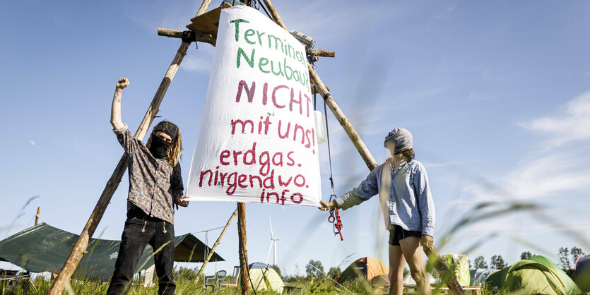 Links und rechts neben einem Gestell mit einem Protestbanner stehen zwei Menschen auf einer sommerlichen Wiese. Die Person links reckt die Faust in die Höhe. Auf dem Banner steht: "Terminal Neubau nicht mit uns. Erdgas.nirgendwo.info"