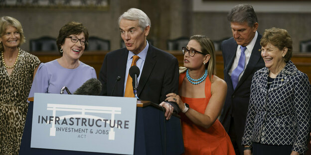 Männliche und weibliche Senatoren stehen um ein Pult mit der Aufschrift "Infrastruktur"