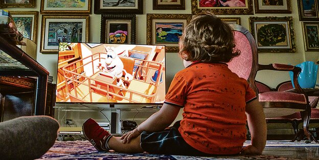 Ein Kind sitzt auf dem Boden, der Rücken zur Kamera gedreht, das Gesicht zum Fernseher. Im Hintergrund hängen Bilderrahmen